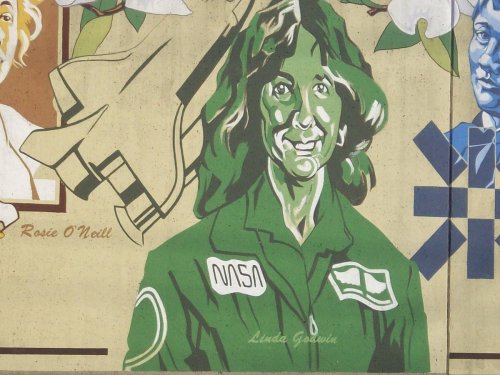 Mural of a woman in a green NASA jumpsuit, smiling, with a space shuttle and flowers in the background.