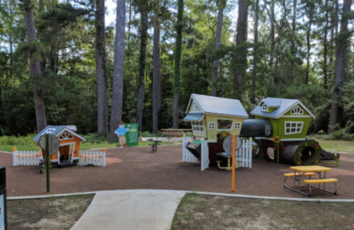 Colorful playground with house-shaped structures, picnic tables, and surrounded by tall trees in a forested area.
