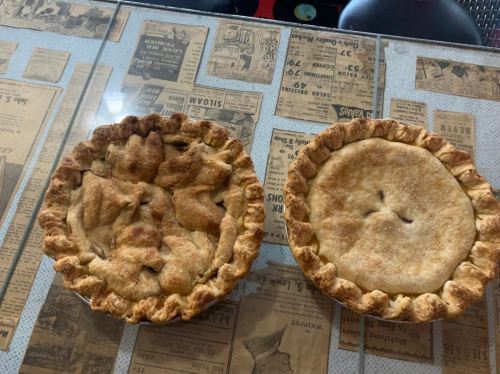 Two freshly baked pies on a table covered with newspaper clippings, one with a lattice top and the other smooth.