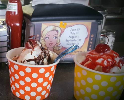 Two ice cream cups with toppings, one brown and one yellow, in front of a diner schedule featuring a retro illustration.