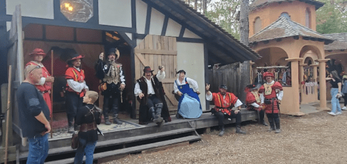 A group of people in medieval costumes poses outside a rustic building at a festival, with vendors in the background.