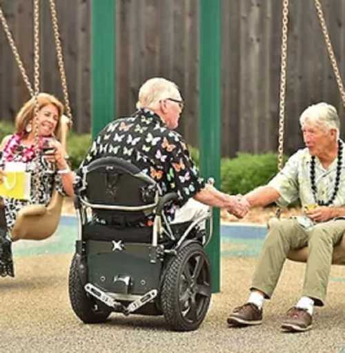 Two elderly men sit on a playground, one in a wheelchair, while a woman swings nearby, all enjoying a sunny day.