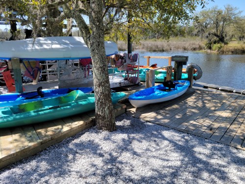 A dock with colorful kayaks, surrounded by trees and a calm river, with people relaxing in the background.