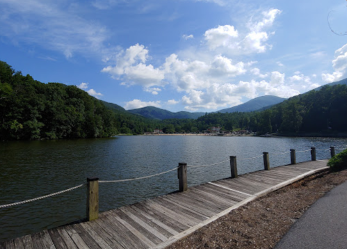 Scenic view of a calm lake surrounded by green mountains and a blue sky with fluffy clouds. Wooden walkway in the foreground.