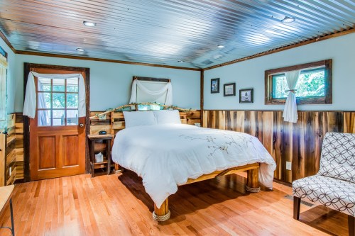 Cozy bedroom with a wooden bed, rustic walls, and large windows, featuring a chair and natural light.