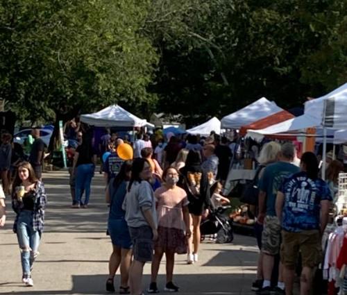 A bustling outdoor market with people browsing stalls and tents under trees on a sunny day.
