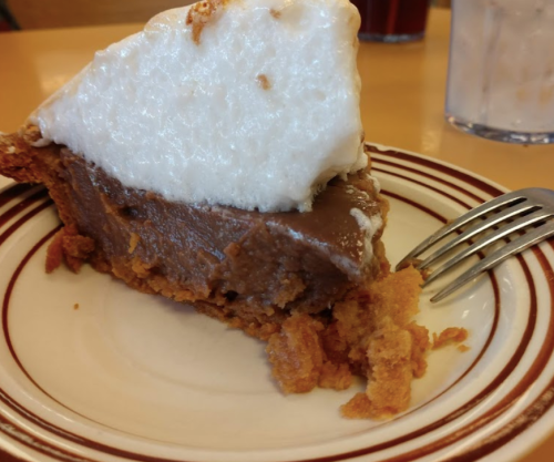 A slice of chocolate pie with a fluffy meringue topping on a striped plate, accompanied by a fork.