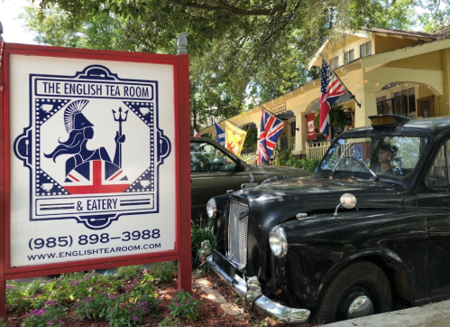 A vintage car parked beside a sign for The English Tea Room & Eatery, adorned with flags and greenery.
