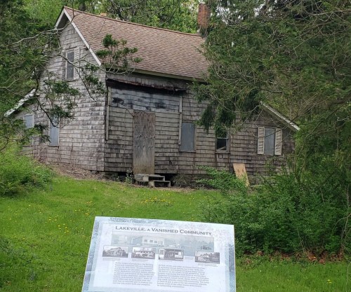 An old, weathered house surrounded by greenery, with an informational sign in the foreground about Lakeville's vanished community.