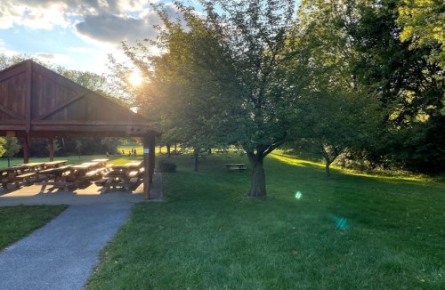 A sunlit park scene featuring a wooden pavilion, grassy area, and trees under a partly cloudy sky.