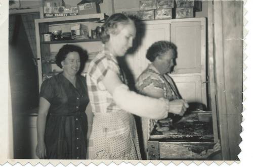 Three women in a kitchen, one cooking at a stove, with shelves of food in the background. Black and white photo.