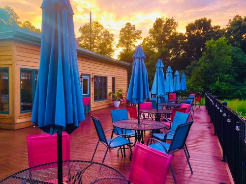 A vibrant outdoor patio with blue umbrellas and colorful chairs, set against a sunset sky and lush greenery.