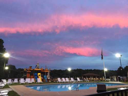 A swimming pool at sunset, with colorful clouds and a water slide in the background, surrounded by lounge chairs.