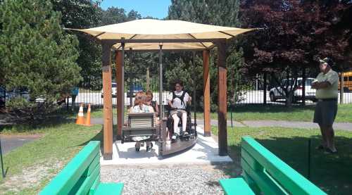 Two people sit on a swing under a shaded structure in a park, surrounded by greenery and a gravel path.