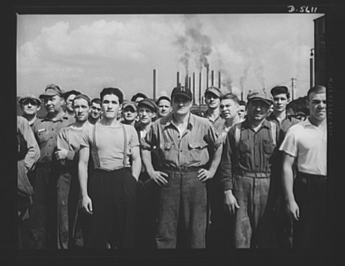 A group of workers stands together, wearing uniforms, with industrial smokestacks in the background under a cloudy sky.