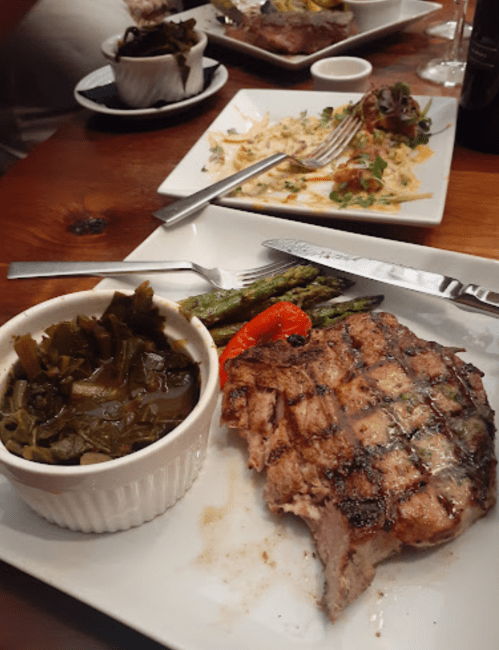 A grilled steak with sides of collard greens and asparagus on a white plate, with another dish in the background.