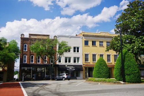 A charming street scene featuring historic buildings, lush trees, and a clear blue sky with fluffy clouds.