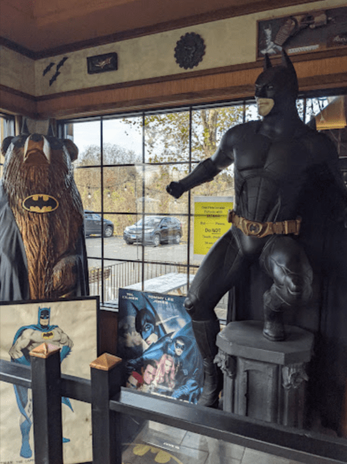 A Batman statue stands next to a bear figure, with Batman-themed artwork displayed nearby in a cozy setting.