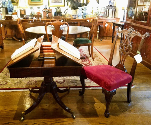A wooden writing desk with open books and a decorative chair in an elegant antique shop setting.