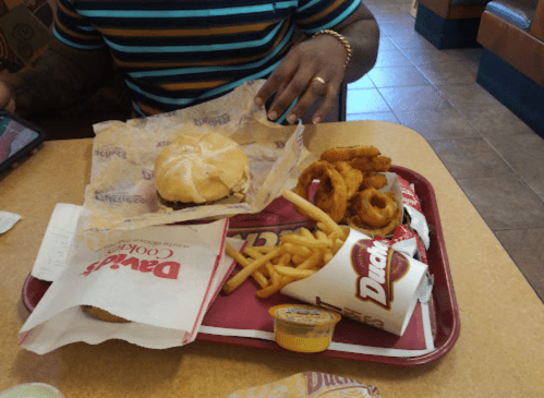 A tray with a burger, onion rings, fries, and dipping sauce, placed on a table in a fast-food restaurant.