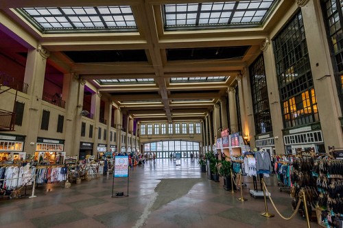 Interior of a spacious, historic building with high ceilings, large windows, and vendor displays along the sides.