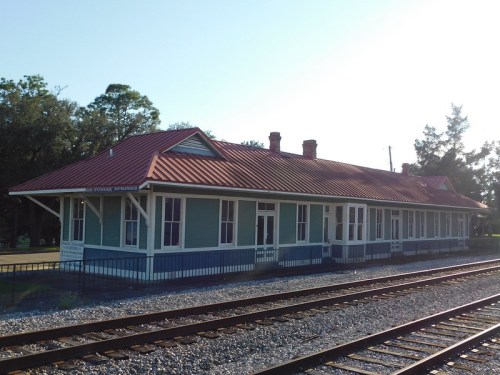 Historic train station with a red metal roof, blue siding, and white trim, situated beside railroad tracks.
