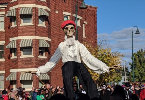 A large skeleton puppet in a white shirt and red hat stands among a crowd near a brick building.