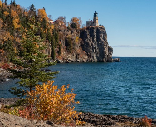 A scenic view of a lighthouse on a cliff, surrounded by autumn foliage and a calm blue lake under a clear sky.