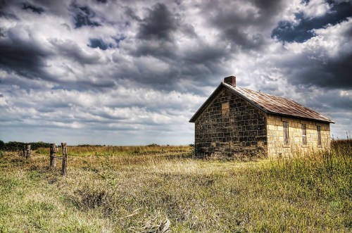 A weathered stone house stands in a grassy field under a dramatic sky with clouds. A fence is visible nearby.