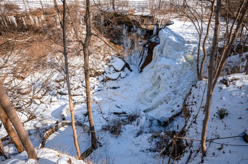 A frozen waterfall surrounded by snow-covered trees and rocky terrain in a winter landscape.