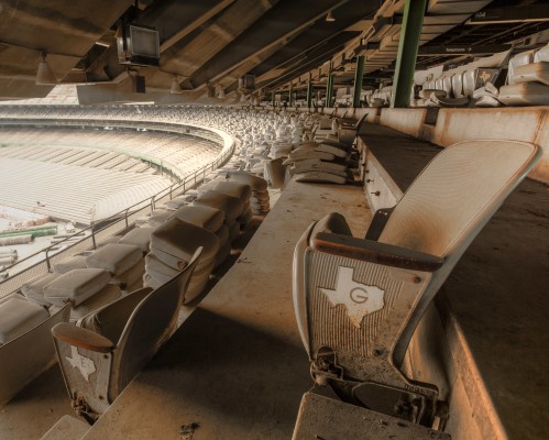 Abandoned stadium seats with a Texas logo, showing dust and neglect in a large, empty arena.