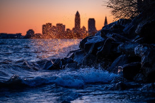 Waves crash against rocky shore at sunset, with a city skyline silhouetted in the background.
