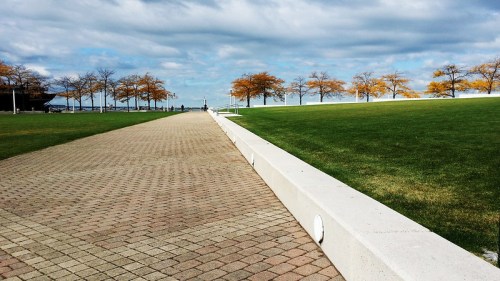 A paved pathway leads through a park with green grass and trees, under a cloudy sky.