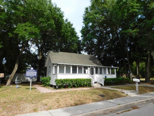 A charming white house with a porch, surrounded by trees and greenery, located on a quiet street.