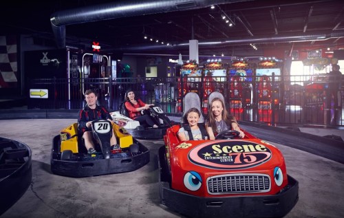 Two go-karts on a track with four kids smiling, surrounded by arcade games in a fun indoor setting.