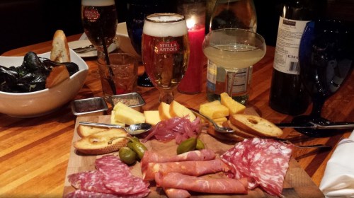 A wooden platter with assorted meats, cheeses, olives, and bread, accompanied by drinks and a bowl of mussels.