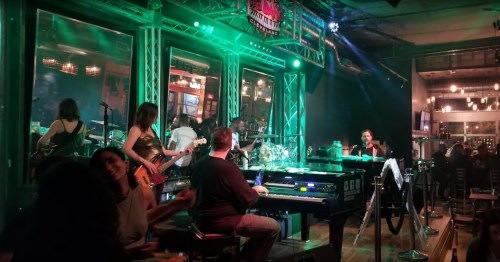 A lively band performs on stage in a dimly lit venue, with green lights and an audience enjoying the music.