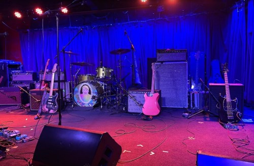 A stage setup with guitars, amplifiers, and a drum kit under blue lighting, ready for a live music performance.