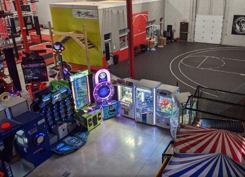 A colorful arcade area with various games and machines, set in a spacious indoor facility with a sports court.