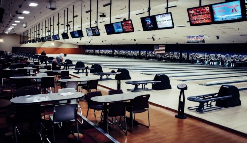 A spacious bowling alley with empty lanes, tables, and screens displaying scores in a well-lit environment.