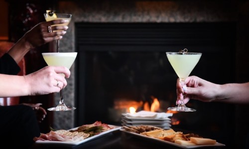 Two hands hold cocktails in front of a cozy fireplace, with a platter of cheese and charcuterie nearby.