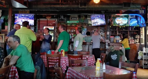 A lively bar scene with patrons in green shirts, watching sports on TV, and enjoying drinks at checkered tables.