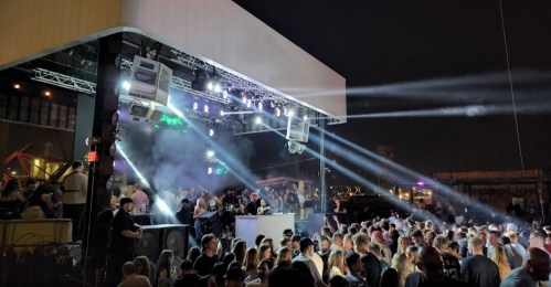 A lively outdoor party scene with a crowd dancing under colorful lights and smoke effects at night.