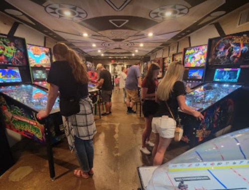 A lively arcade with people playing pinball machines in a colorful, retro-themed environment.