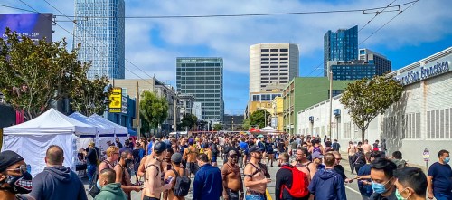 A bustling street scene with a large crowd, tents, and city buildings in the background on a sunny day.