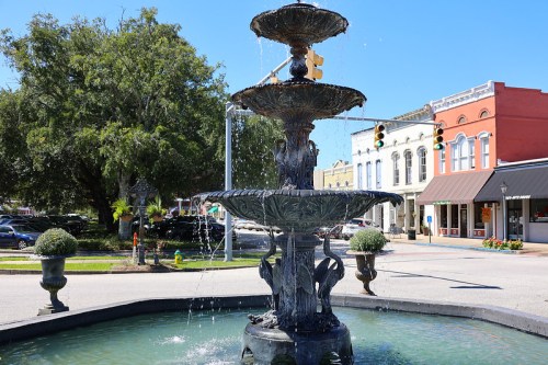 A decorative three-tiered fountain in a town square, surrounded by greenery and historic buildings under a clear blue sky.