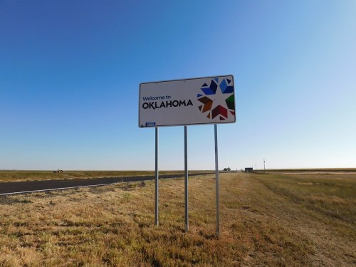 Welcome sign for Oklahoma against a clear blue sky, with open fields and a distant road.