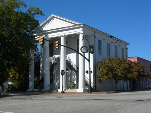 Historic white building with columns, surrounded by trees, at a street intersection with traffic lights and wreaths.