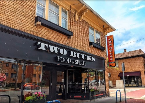 Exterior of "Two Bucks Food & Spirit," a brick building with a sign, featuring large windows and a welcoming entrance.
