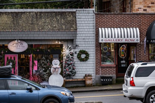 A festive street scene featuring shops decorated for the holidays, including a snowman and wreaths, with cars parked nearby.